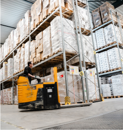 Ein Gabelstapler fährt durch ein Lager mit hohen Regalen voller Paletten.