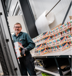 Mann in Arbeitskleidung, der eine Kiste hält, vor einem Regal mit bunten Verpackungen in einer Lagerhalle.